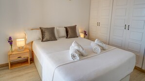 Een netjes opgemaakt bed met witte lakens en handdoeken, een nachtkastje met een lamp en bloemen, en een garderobe met meerdere deuren.