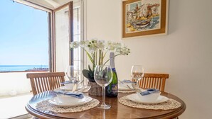 Een ronde houten eettafel met witte borden, blauwe servetten en een fles wijn. Er zijn twee stoelen, een potplant en een lijst met een schilderij aan de muur.