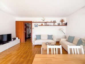 Sala de estar com televisão, sofá, mesa de jantar e cadeiras. O chão é de madeira e há uma porta que dá para outro cômodo.