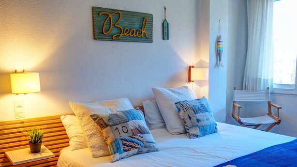 Ein ordentlich eingerichtetes Hotelzimmer mit einem Bett, zwei Kissen, einem Nachttisch mit Lampe, einer kleinen Pflanze und einer Wanddekoration.