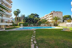 A swimming pool surrounded by a well-maintained lawn and palm trees, with modern buildings in the background. - Clarimar Apartment by Hello Homes Sitges (Sitges)