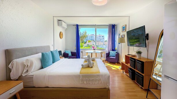 A hotel room with a bed, a TV, a desk, a chair, and a view of buildings. - The Blue Island Loft by Hello Homes Sitges (Sitges)