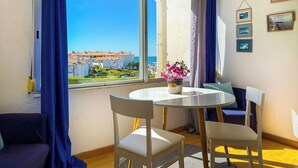 A room with a round table, two chairs, a sofa, a window with curtains, and a view of buildings and the sea. - The Blue Island Loft by Hello Homes Sitges (Sitges)