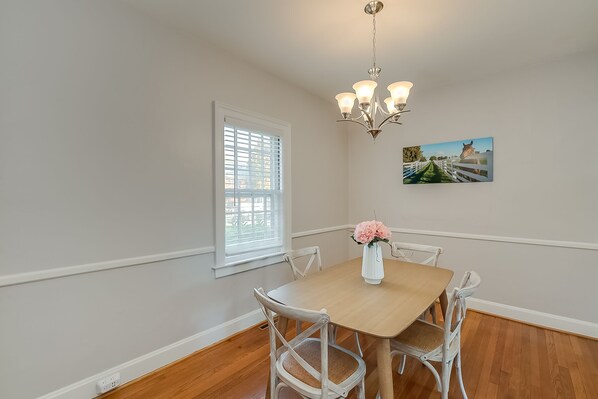 A dining area with a wooden table, white chairs, a chandelier, a window with blinds, and a painting on the wall.