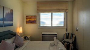 Habitación de hotel con cama, ventana con vista al mar, un cuadro en la pared y una silla.