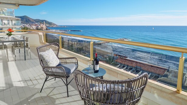 Un balcón con sillas de mimbre, una mesa y una botella de vino, con vistas a un pueblo costero y al mar.