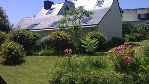 Una casa amb teulada inclinada, diverses claranes i envoltada de vegetació i plantes florides.