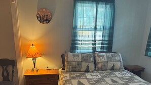 A bedroom with a bed, a nightstand, a lamp, a window with curtains, and a wall clock.
