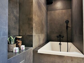 A modern bathroom with a freestanding bathtub, dark tiled walls, and a small shelf with toiletries.