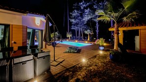 Uma área de piscina com deck de madeira, móveis de exterior e um pequeno edifício com telhado inclinado.