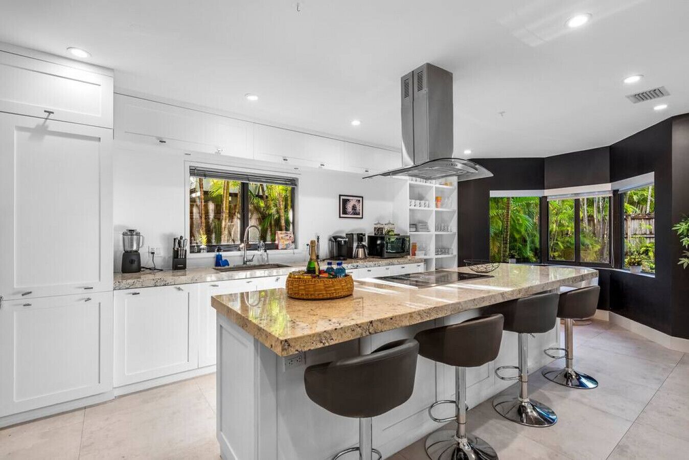 A modern kitchen with white cabinets, a large island with bar stools, a built-in stove, and a large window overlooking greenery.