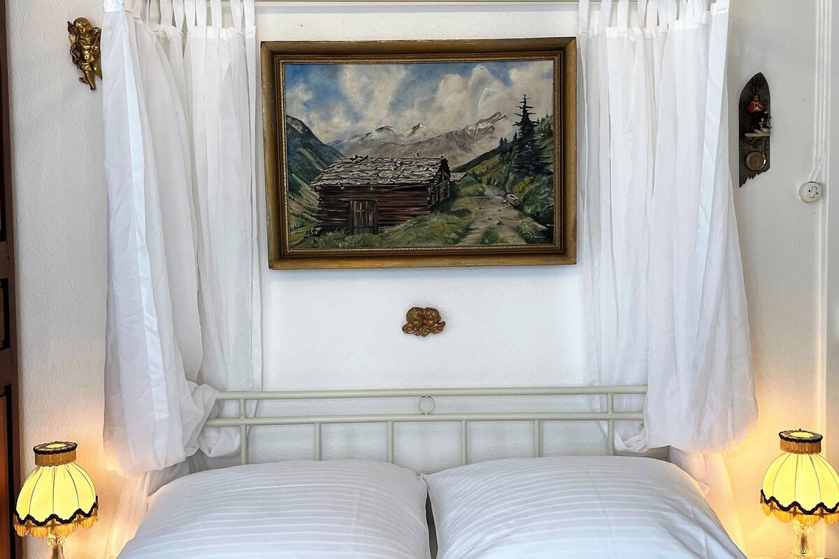 A bedroom with a bed, two bedside lamps, a painting of a mountain scene, and a small decorative angel on the wall.