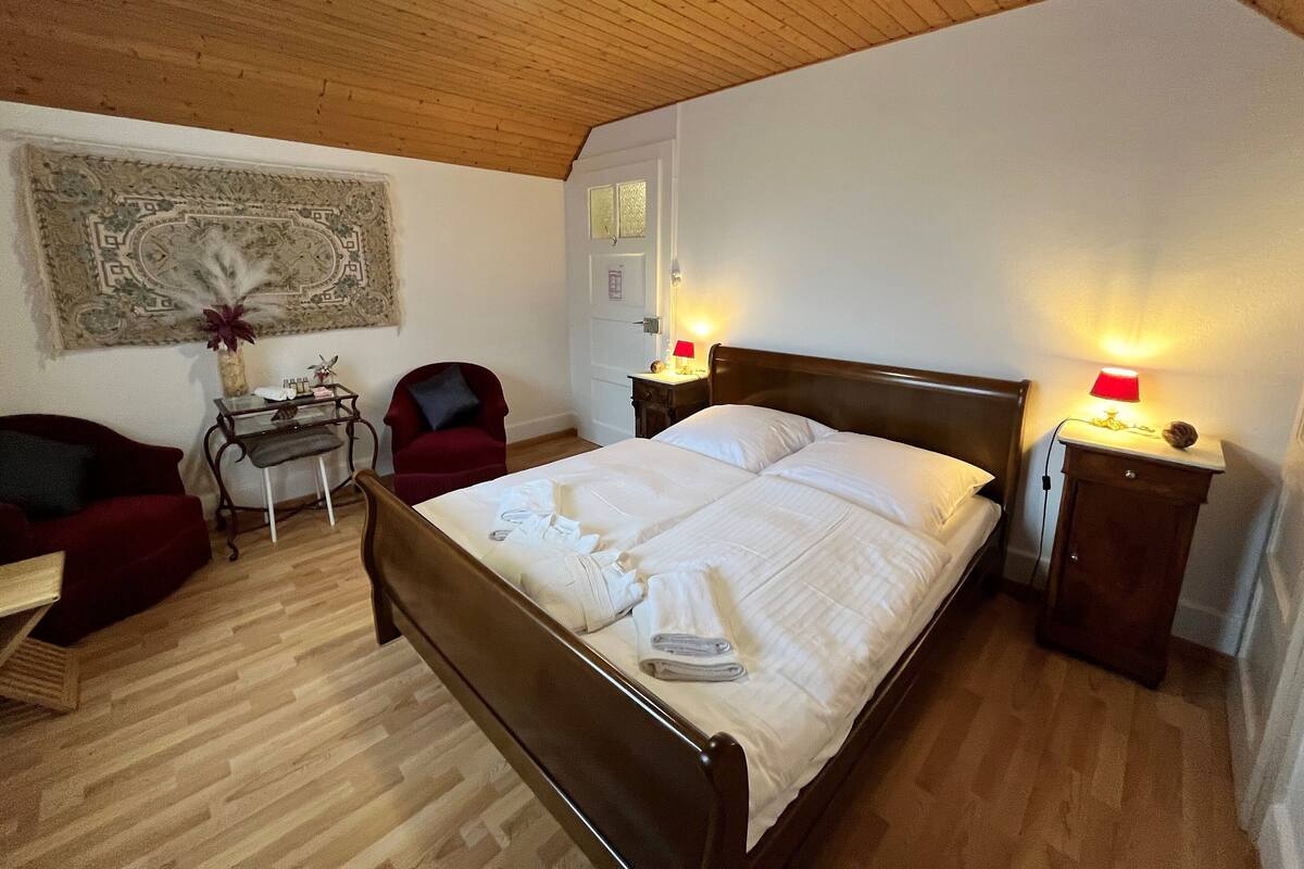 A bedroom with a wooden ceiling, a bed with white linens, a red chair, a small table, and two bedside lamps.