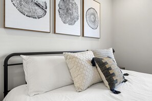 Un lit avec une literie blanche et deux coussins décoratifs, une tête de lit en métal noir et trois reproductions botaniques encadrées accrochées au mur.