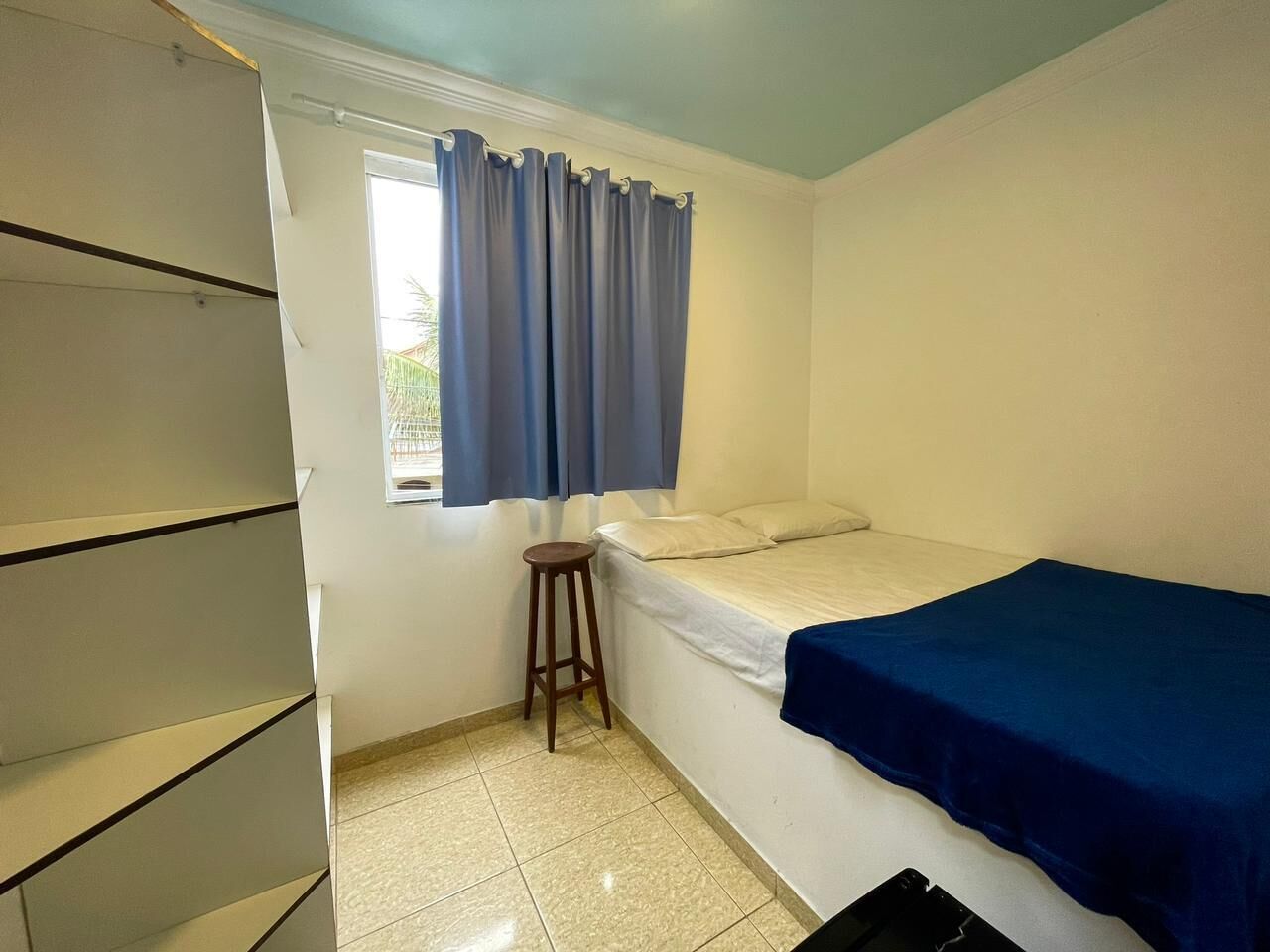 Una habitación pequeña y limpia, con una cama, un taburete y un armario.