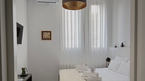 Ein Hotelzimmer mit Bett, Nachttisch mit Lampe, einem Bild an der Wand und einem Fenster mit durchsichtigen Vorhängen.
