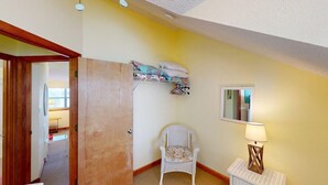 A room with a wooden door, a wicker chair, a small table with a lamp, and a shelf with a pillow. - Affordable Gulf Front Triplex (Port Saint Joe)