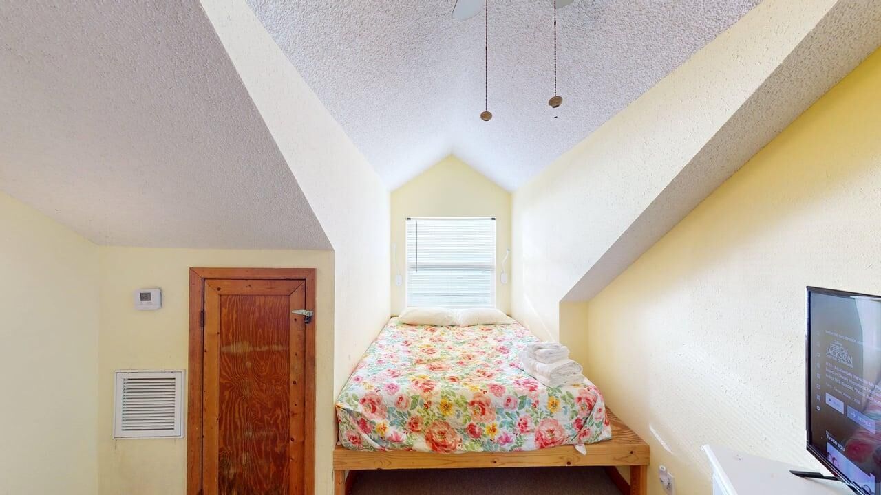 A small, single-bed room with a sloped ceiling, a wooden door, a window with blinds, and a television on a desk.