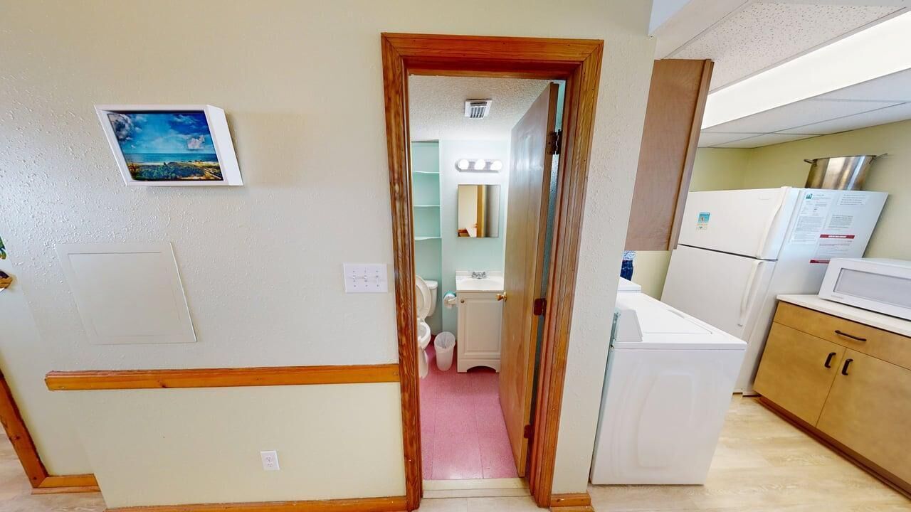 A narrow hallway leading to a bathroom with a toilet, sink, and mirror. There is a white refrigerator and microwave on the right, and a wooden cabinet with a microwave on top.
