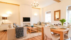 A modern living room with a sofa, coffee table, dining table, and chairs. There is a TV mounted on the wall, a chandelier, and a window with a view. - Feathers, Blythburgh (Halesworth)