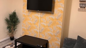 A living room with a flat-screen TV mounted on a yellow patterned wall, a black coffee table, a grey sofa, and a potted plant. - Budget 5-bed Apartment in Barking (Barking)