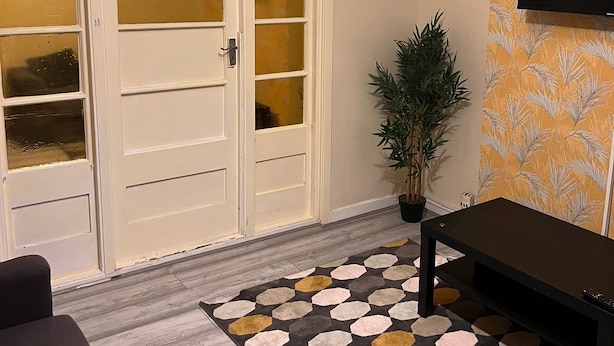 A room with a large sliding glass door, a potted plant, a black bench, and a wall-mounted TV. - Budget 5-bed Apartment in Barking (Barking)