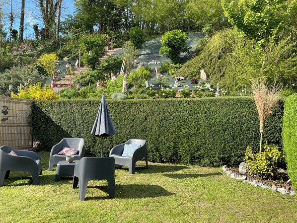 Un jardín bien cuidado con una zona de estar que incluye sillas de plástico y una mesa, rodeado de setos ordenados y diversas plantas.