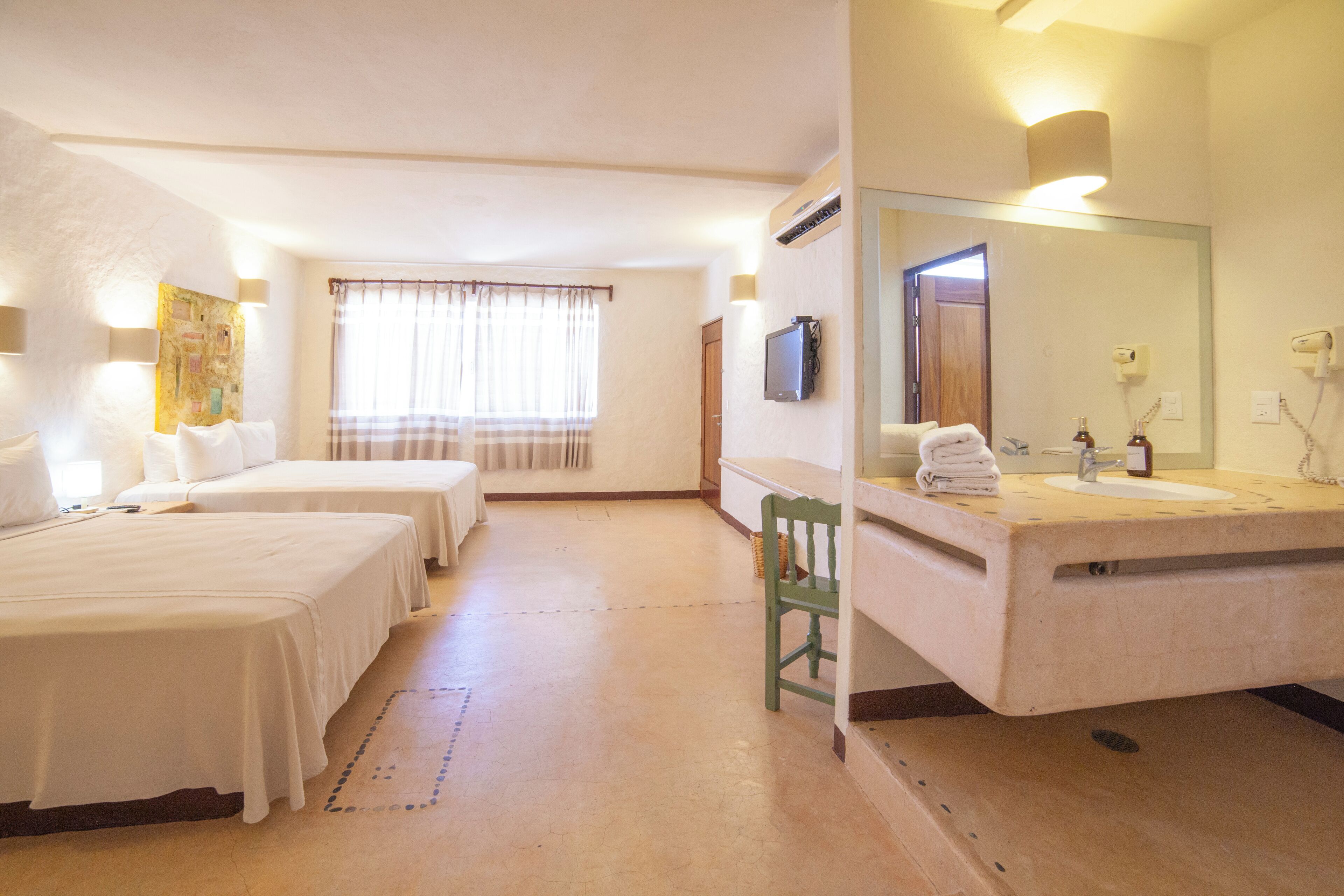 A hotel room with two beds, a bathroom with a sink and mirror, and a window with curtains.
