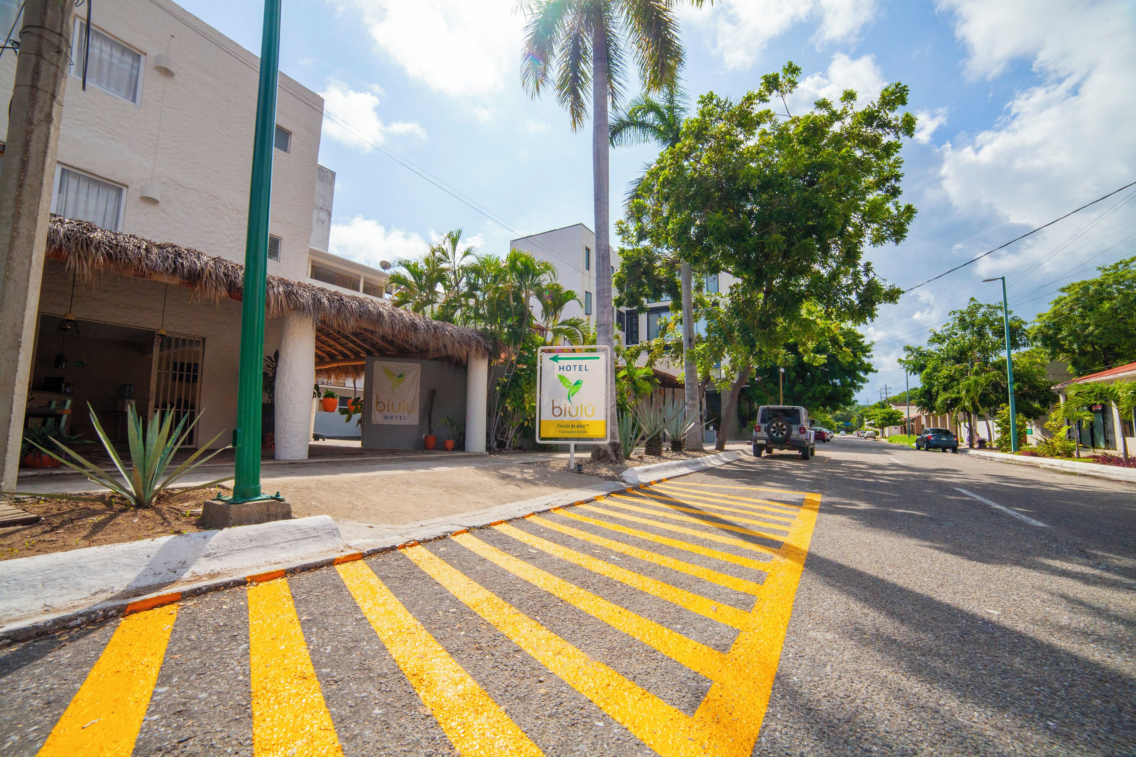 Un hotel con un cartel que indica la cercanía de otro hotel, una calle con marcas direccionales amarillas y palmeras.