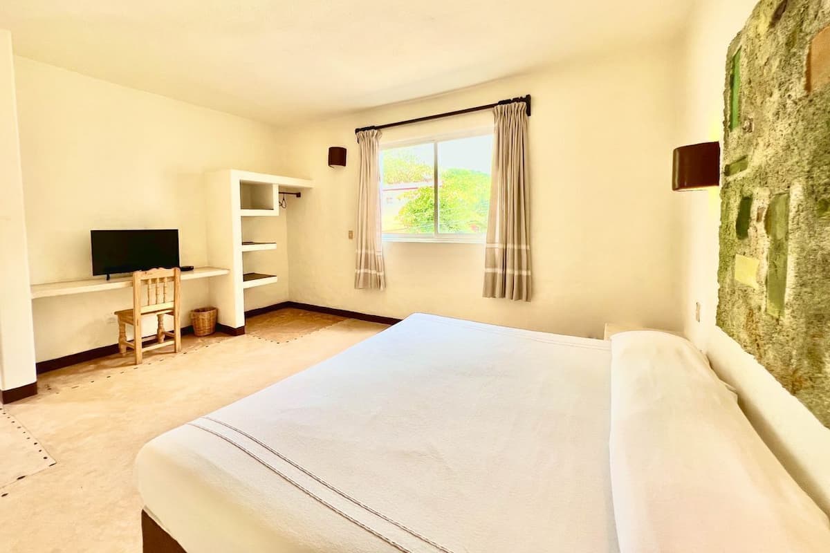 A bedroom with a bed, a TV, a shelf, a window with curtains, and a stone wall.