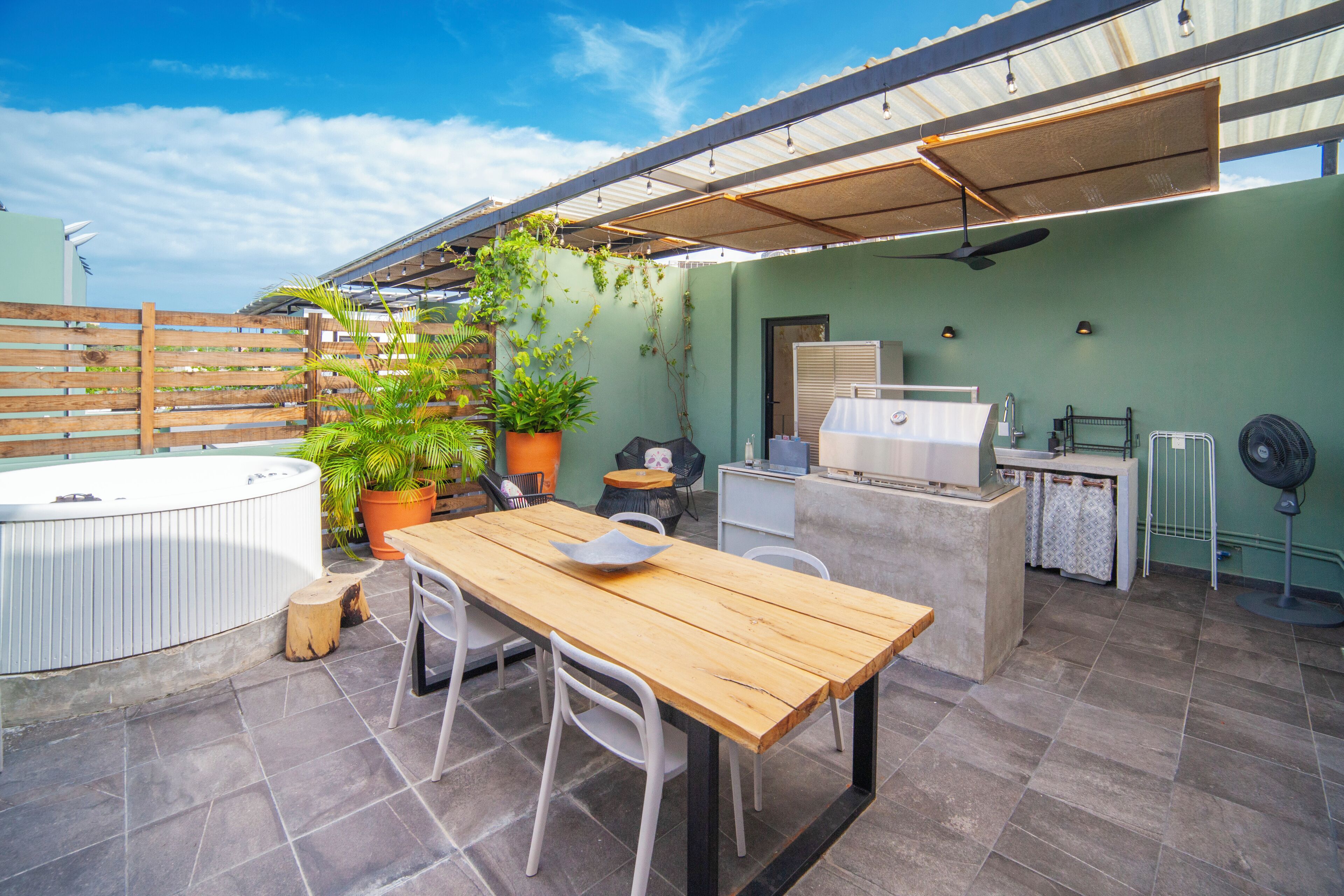 An outdoor patio with a wooden table and chairs, a hot tub, potted plants, and a grill area.