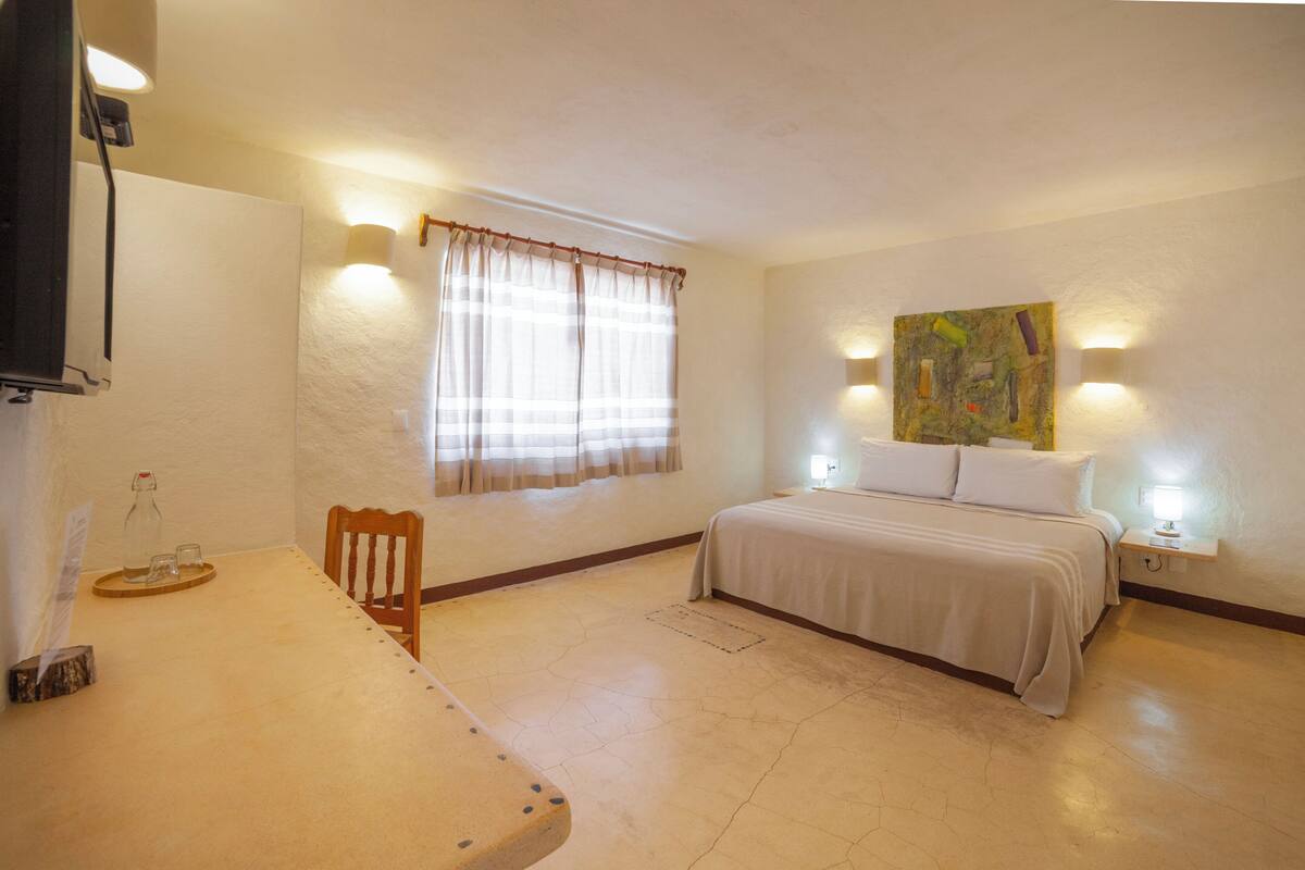 A bedroom with a bed, a chair, a table with a bottle and glasses, and a painting on the wall.