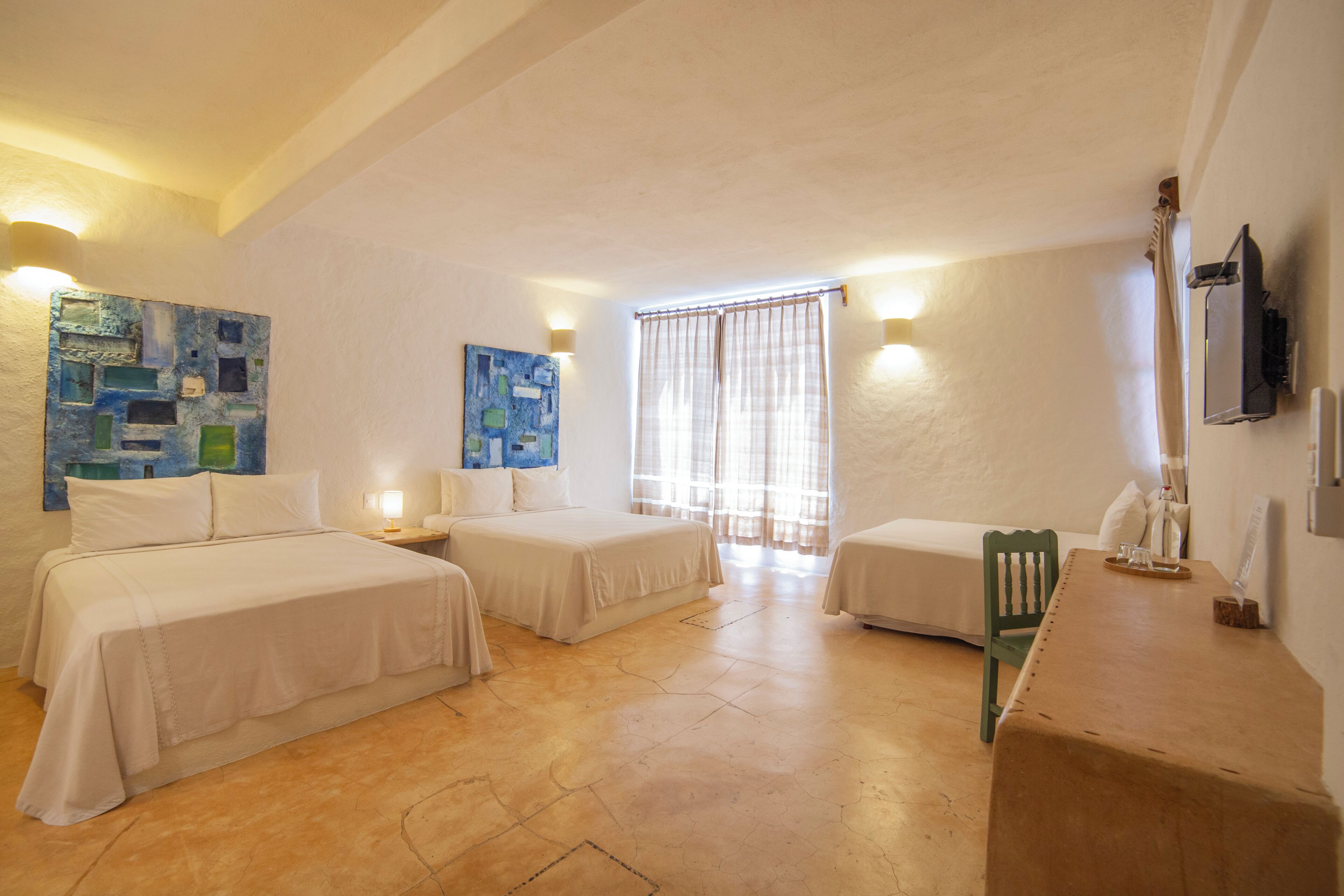 A room with two beds, a small table, and a chair. There are two paintings on the walls and a window with curtains.
