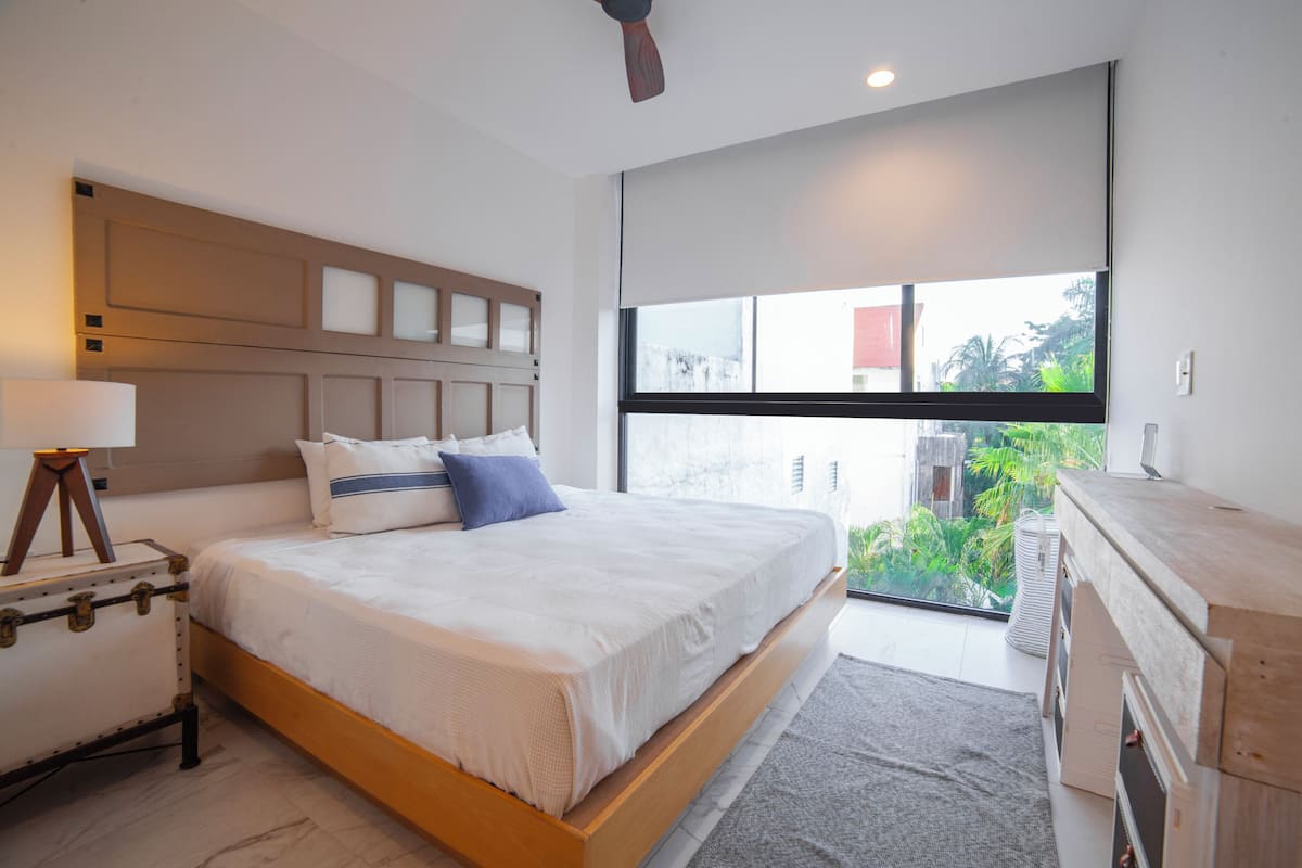 A modern bedroom with a large bed, a bedside table with a lamp, a wooden headboard, and a view of greenery through a large window.