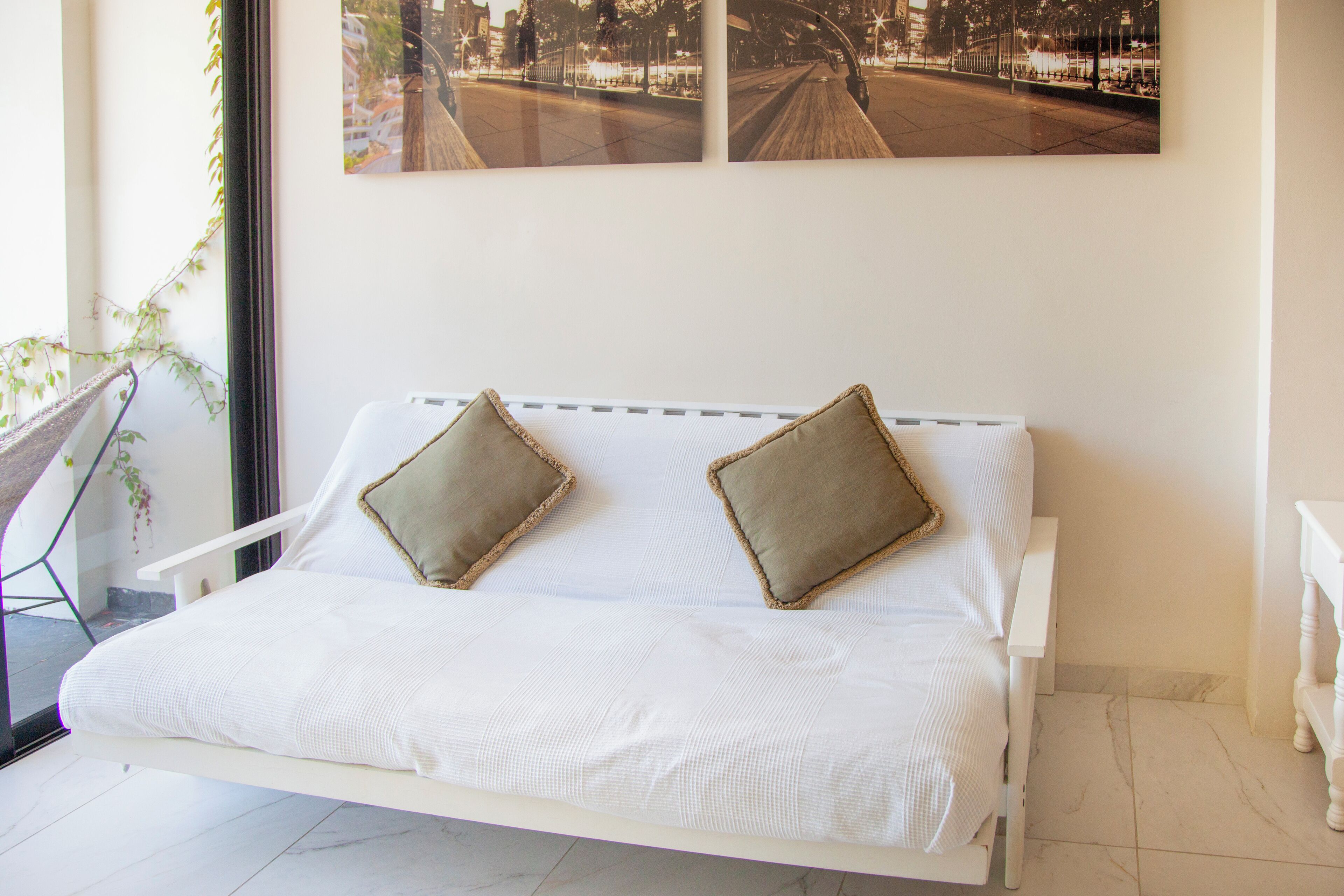 A white bed with two pillows, a window with a view of a street, and a plant beside the window.