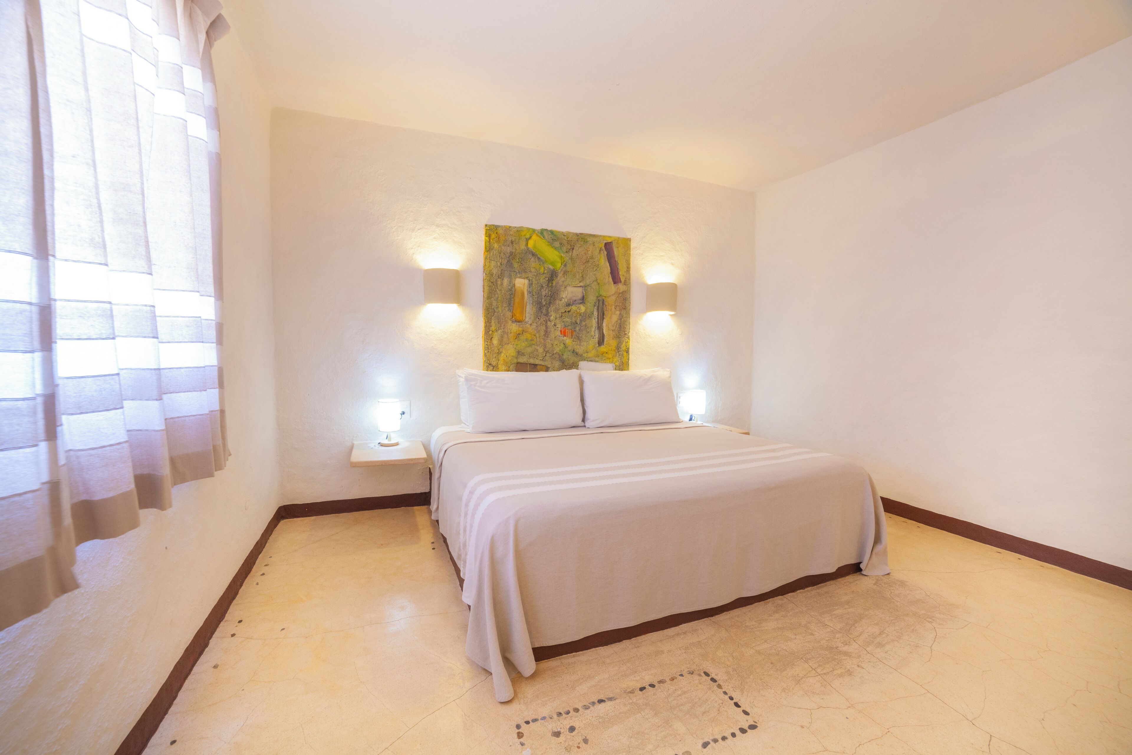 A bedroom with a bed, two bedside lamps, a wall-mounted painting, and a window with curtains.