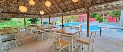 Uma área de refeições ao ar livre coberta, com mesas e cadeiras, uma piscina ao fundo e uma estrutura com telhado de palha.