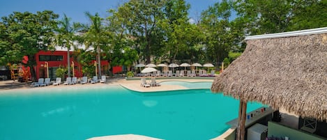 A swimming pool with lounge chairs and umbrellas, surrounded by trees and a building with a thatched roof.