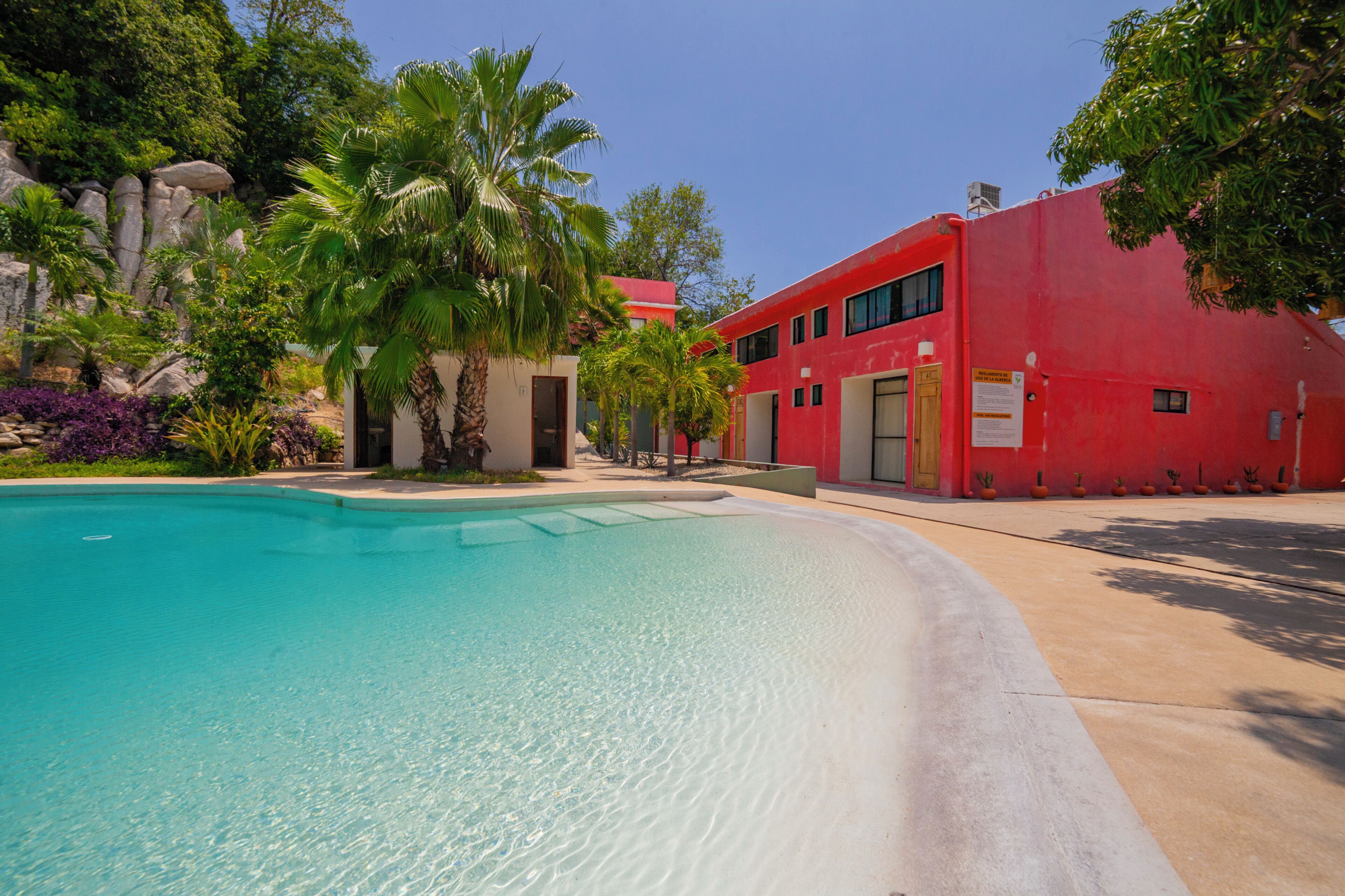 Una piscina con agua cristalina, rodeada de vegetación tropical y un edificio rojo con varias ventanas.