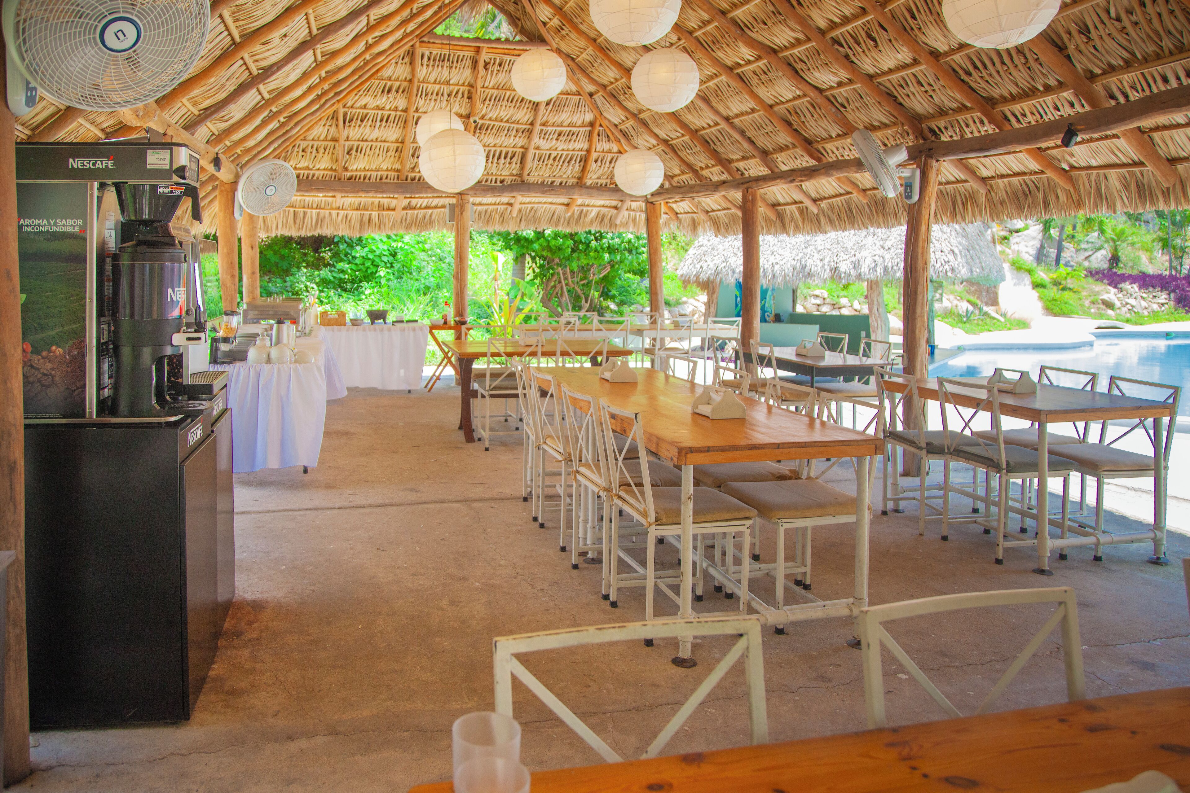 Un área de comedor al aire libre con mesas y sillas, una cafetera y una piscina al fondo.