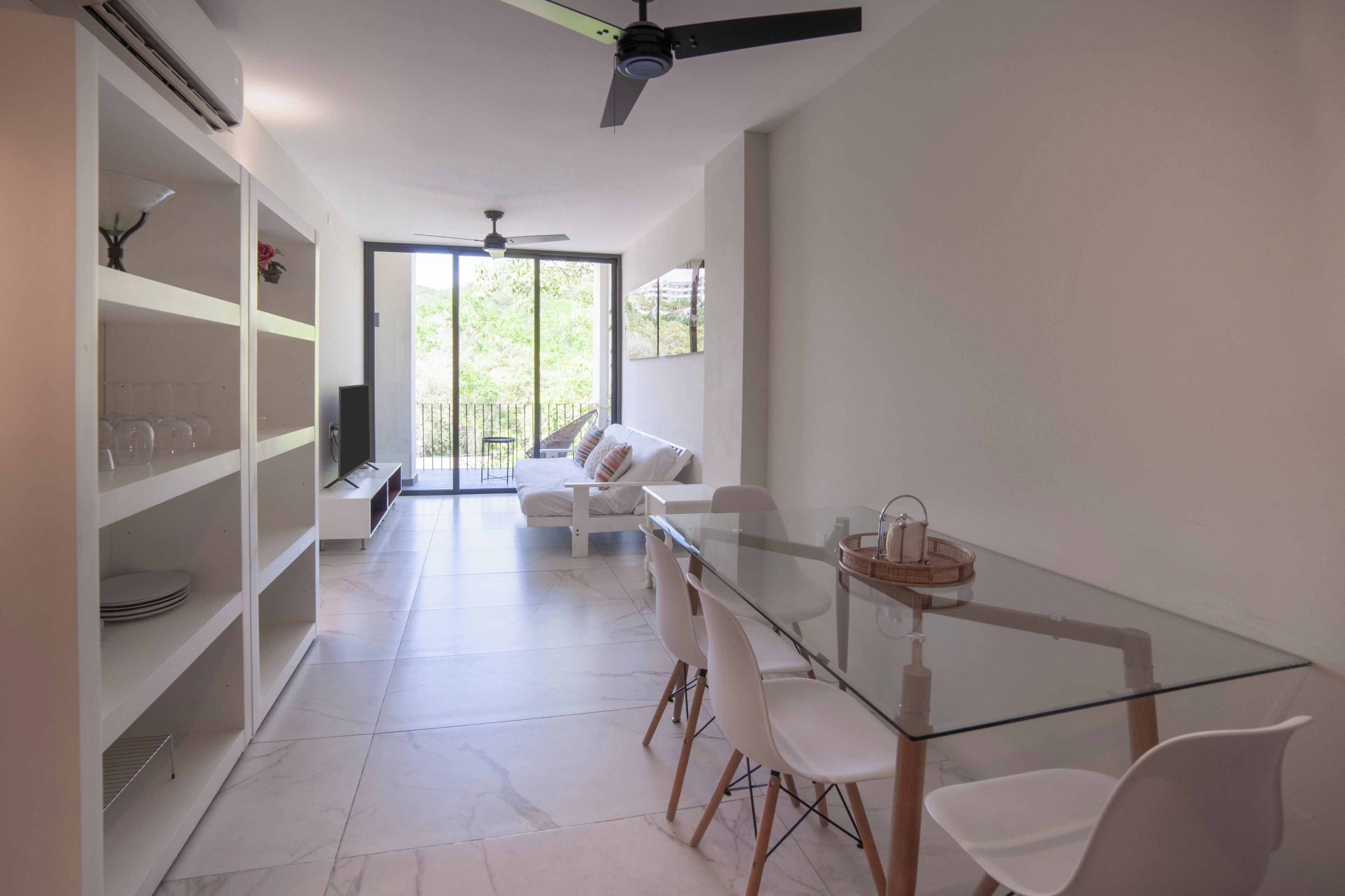 A modern living room with a glass dining table, white chairs, a sofa, and built-in shelving.