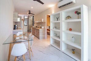A modern kitchen with a glass dining table, white chairs, and a white shelving unit with decorative items. - HOTEL BIULÚ (Santa María Huatulco)