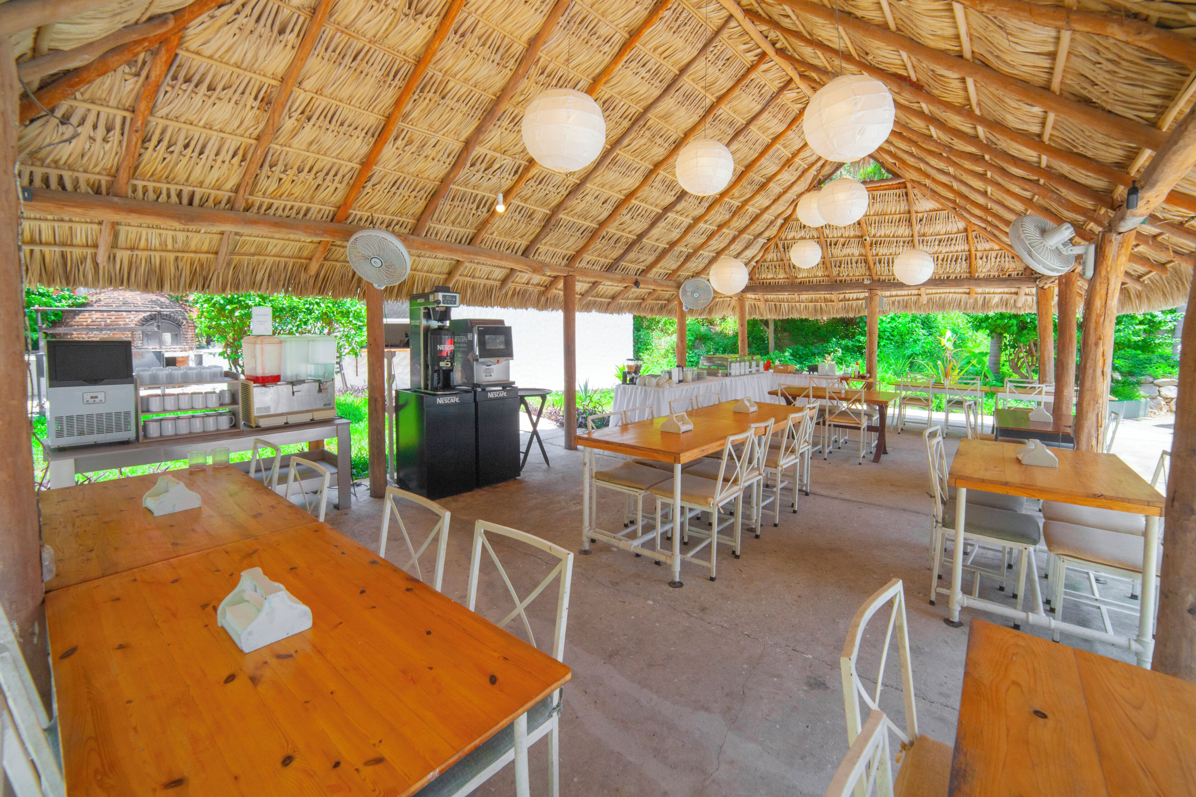 Un área de comedor al aire libre con mesas y sillas de madera, techo de paja y una estación de parrillada.