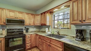 Una cocina con armarios de madera, microondas de acero inoxidable, horno doble, lavavajillas, encimeras de granito y ventana con vista a árboles.