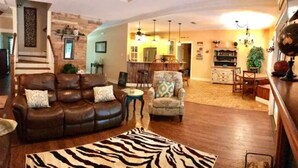 Ein Wohnzimmer mit einem braunen Ledersofa, einem gemusterten Sessel und einem Zebra-Teppich. Eine Treppe mit Holzbalustrade führt zu einer oberen Ebene.