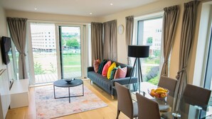 Een moderne woonkamer met een bank, een eettafel en stoelen. Er is een groot raam met gordijnen, een flatscreen televisie aan de muur en een vloerlamp.