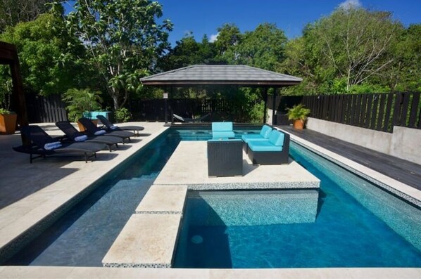 Une piscine rectangulaire avec une terrasse carrelée, entourée de chaises longues et d’un espace salon abrité.
