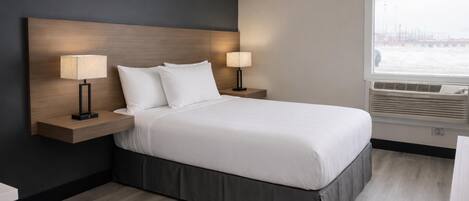 A hotel room with a bed, two bedside lamps, a window with blinds, and a wall-mounted air conditioning unit.