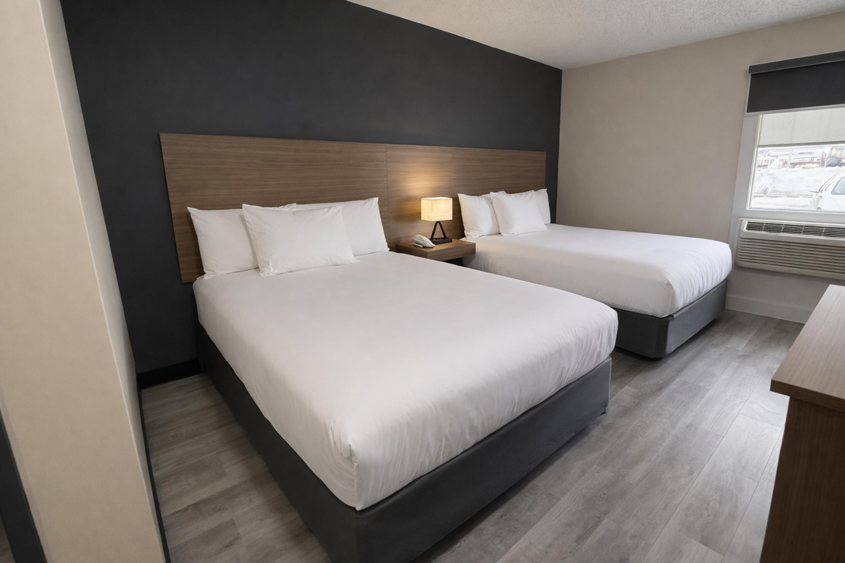 Una habitación de hotel con dos camas, una mesita de noche con lámpara, una ventana con persianas y suelo de madera.
