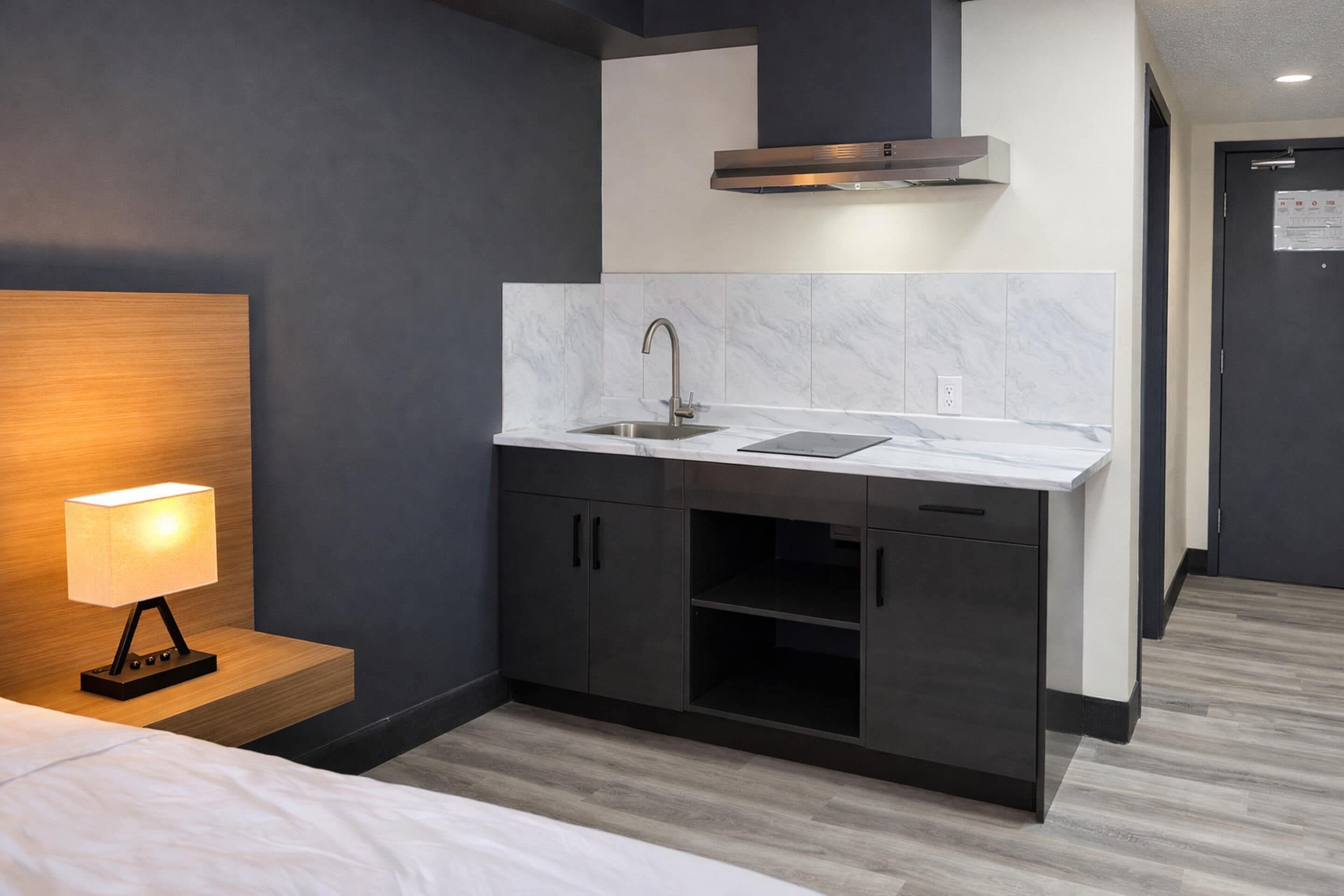 A compact hotel room with a bed, a bedside lamp, a kitchenette with a sink, cabinets, and a stove hood.